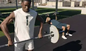 Dos hombres entrenando al aire libre. Uno levanta pesas en banco, el otro sostiene una barra. Esto es que es el crossfit: fuerza y resistencia.