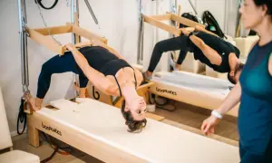 Dos personas practican que es el pilates en equipos de madera, con una mujer en primer plano.
