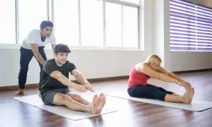 Un instructor ayuda a un hombre a realizar un ejercicio terapéutico de estiramiento sentado. Una mujer también practica el mismo ejercicio.