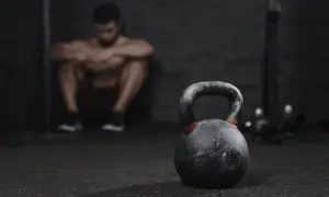 Un hombre musculoso descansa en el gimnasio, con una pesa rusa en primer plano. A veces, es bueno descansar del gym una semana.