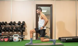Hombre realizando zancadas, un ejercicio de piernas, en un gimnasio con pesas y una cinta de correr al fondo.