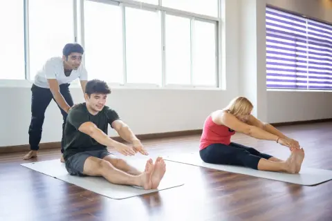 Un instructor ayuda a un hombre a realizar un ejercicio terapéutico de estiramiento sentado. Una mujer también practica el mismo ejercicio.