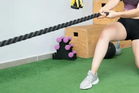 Mujer realizando ejercicios de fisioterapia con una cuerda de entrenamiento. Se ven pesas y cajones de pliometría al fondo.