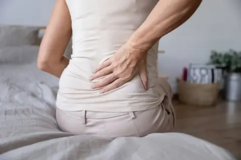 Mujer con dolor de espalda y estómago inflamado, sentada en la cama, con la mano en la zona lumbar.