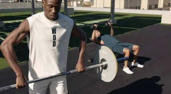 Dos hombres entrenando al aire libre. Uno levanta pesas en banco, el otro sostiene una barra. Esto es que es el crossfit: fuerza y resistencia.