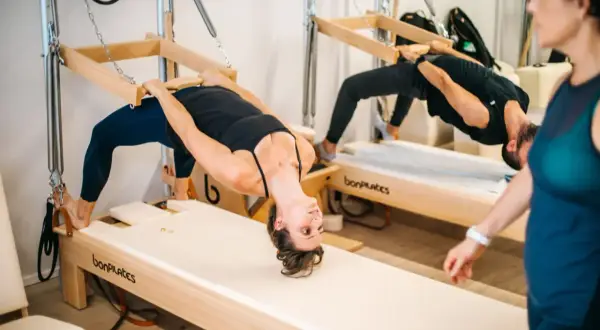 Dos personas practican que es el pilates en equipos de madera, con una mujer en primer plano.