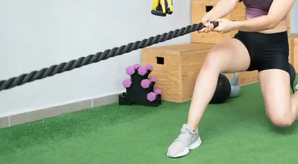 Mujer realizando ejercicios de fisioterapia con una cuerda de entrenamiento. Se ven pesas y cajones de pliometría al fondo.