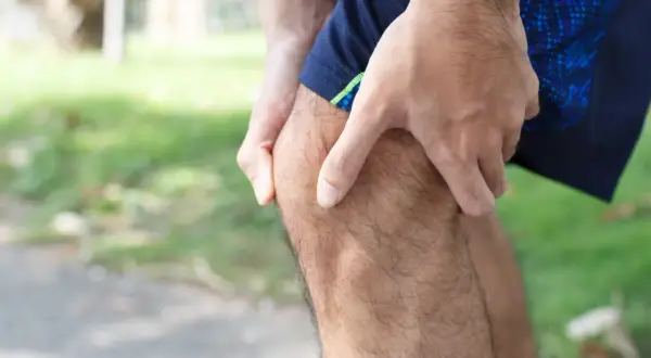 Hombre con dolor parte trasera rodilla al caminar. Sostiene su rodilla con la mano, con ropa deportiva azul.
