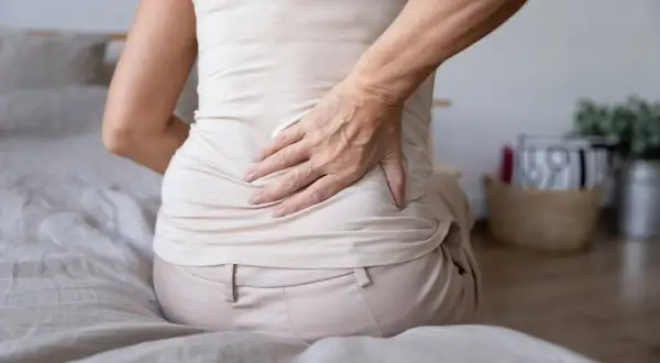 Mujer con dolor de espalda y estómago inflamado, sentada en la cama, con la mano en la zona lumbar.