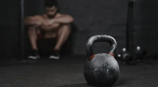 Un hombre musculoso descansa en el gimnasio, con una pesa rusa en primer plano. A veces, es bueno descansar del gym una semana.