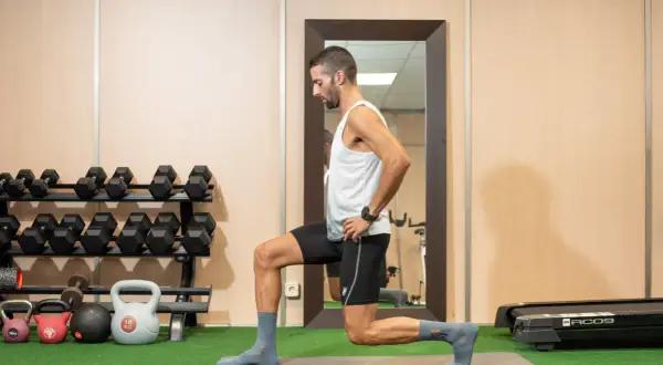 Hombre realizando zancadas, un ejercicio de piernas, en un gimnasio con pesas y una cinta de correr al fondo.