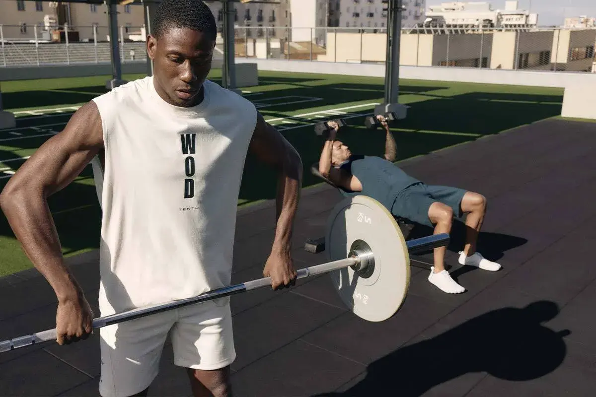Dos hombres entrenando al aire libre. Uno levanta pesas en banco, el otro sostiene una barra. Esto es que es el crossfit: fuerza y resistencia.