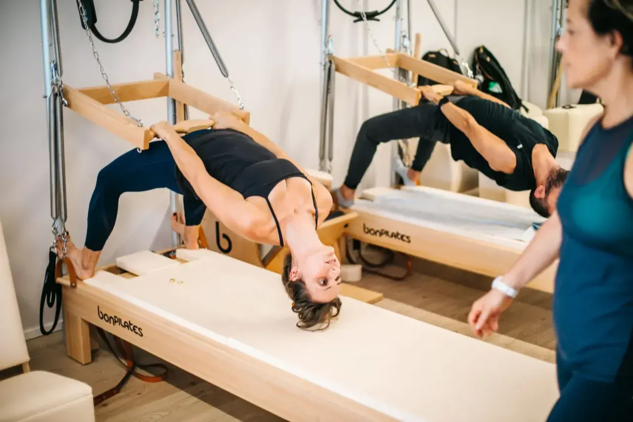 Dos personas practican que es el pilates en equipos de madera, con una mujer en primer plano.