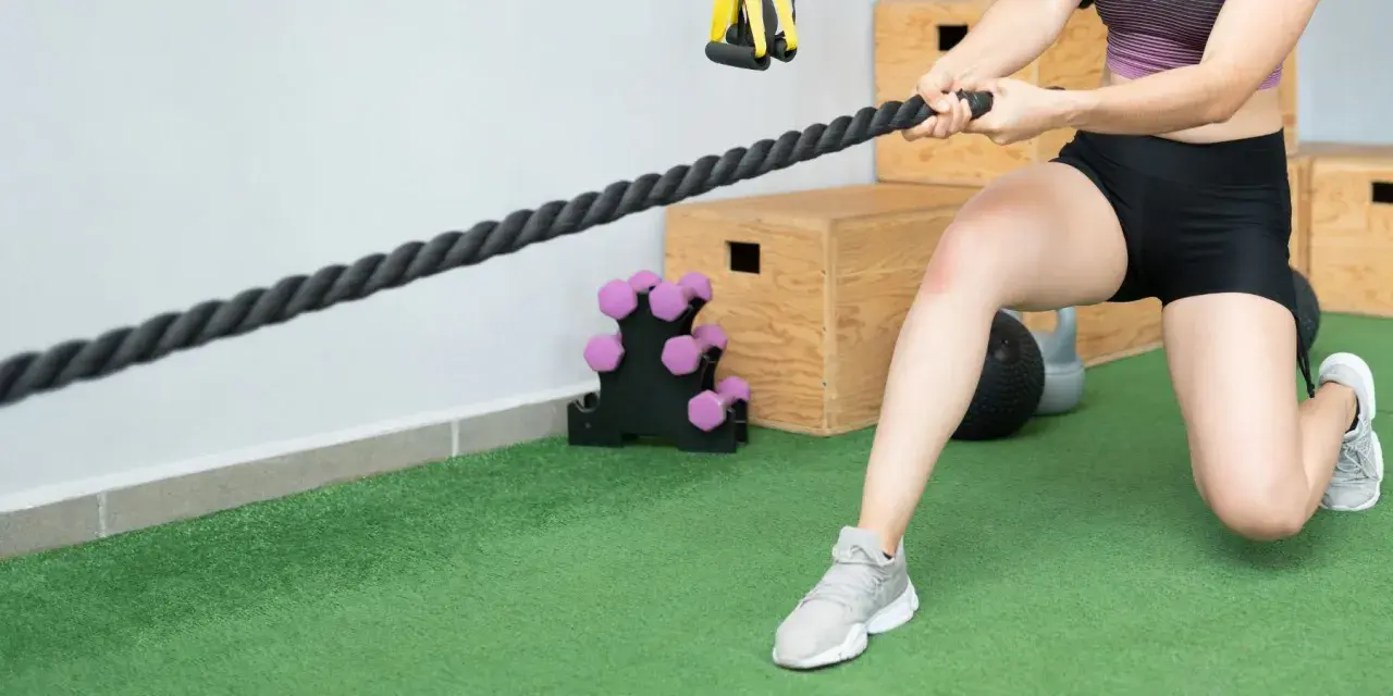 Mujer realizando ejercicios de fisioterapia con una cuerda de entrenamiento. Se ven pesas y cajones de pliometría al fondo.