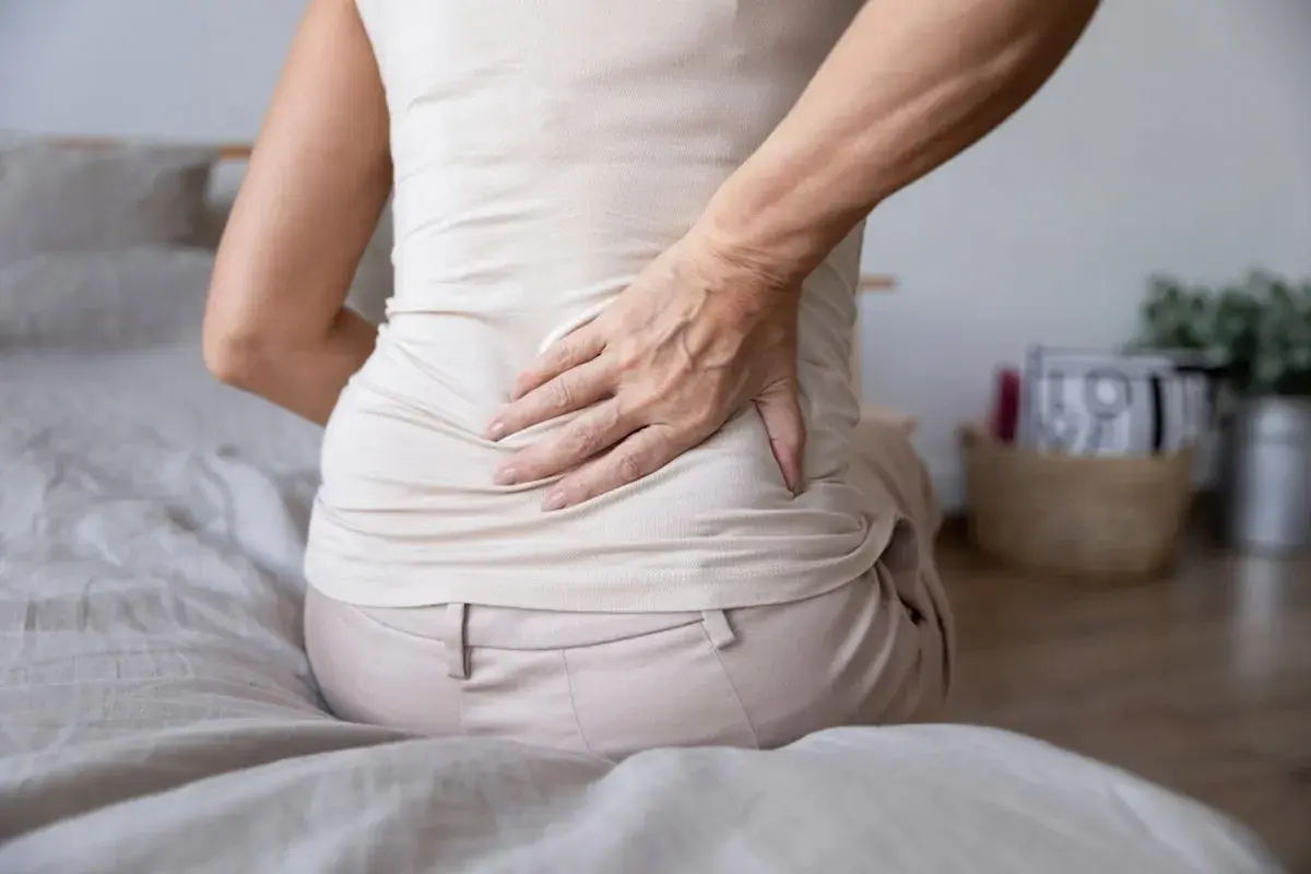 Mujer con dolor de espalda y estómago inflamado, sentada en la cama, con la mano en la zona lumbar.