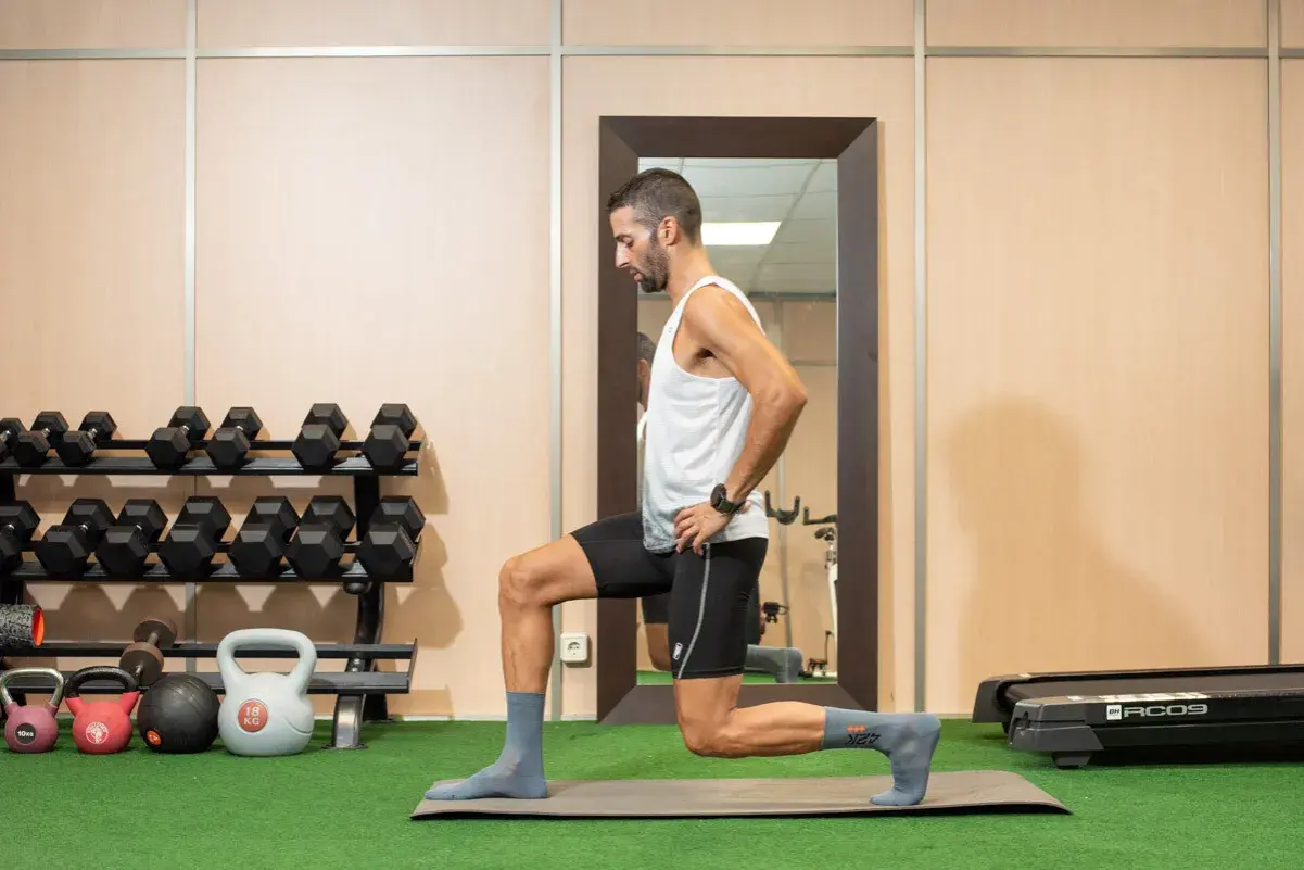 Hombre realizando zancadas, un ejercicio de piernas, en un gimnasio con pesas y una cinta de correr al fondo.