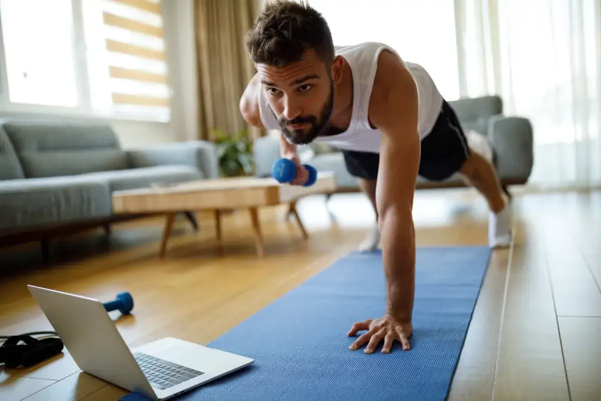 Hombre haciendo ejercicio en casa con mancuernas y port&aacute;til. A veces, es bueno descansar del gym una semana.
