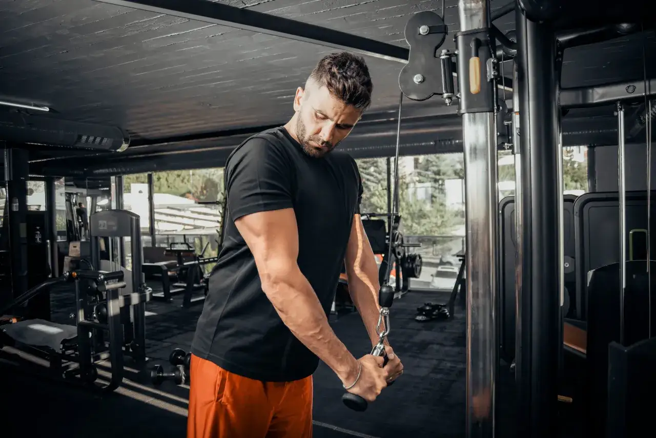 Hombre concentrado realizando ejercicios tr&iacute;ceps en m&aacute;quina de poleas en un gimnasio moderno.
