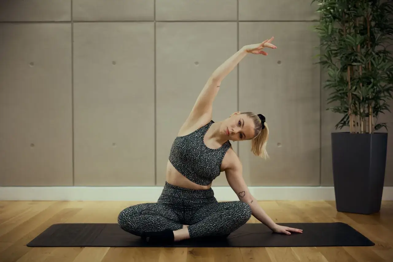 Mujer rubia en ropa deportiva realiza estiramiento lateral sentada en esterilla. Esto es que es el pilates: movimiento y control.