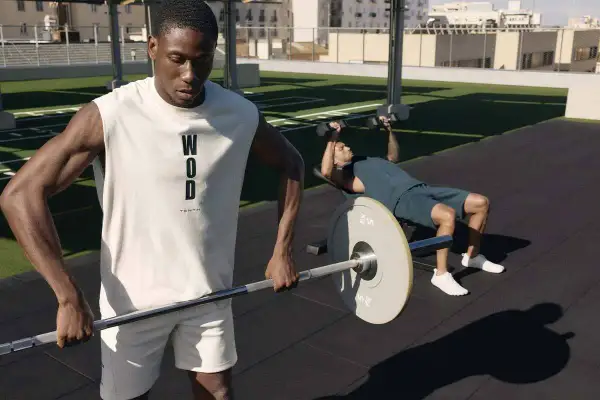 Dos hombres entrenando al aire libre. Uno levanta pesas en banco, el otro sostiene una barra. Esto es que es el crossfit: fuerza y resistencia.