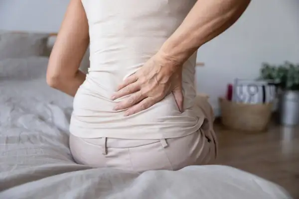 Mujer con dolor de espalda y estómago inflamado, sentada en la cama, con la mano en la zona lumbar.