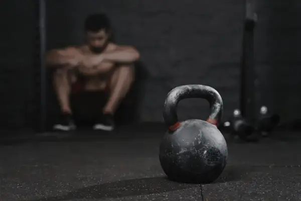 Un hombre musculoso descansa en el gimnasio, con una pesa rusa en primer plano. A veces, es bueno descansar del gym una semana.