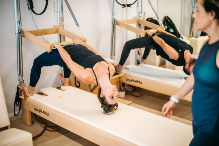 Dos personas practican que es el pilates en equipos de madera, con una mujer en primer plano.